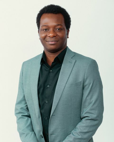 Dapo Bankole smiling, wearing a sage-green blazer over a black shirt, hands clasped, against a light studio background.