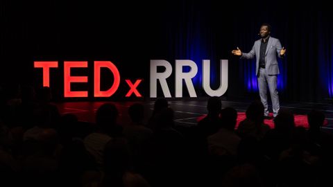 Dapo Bankole speaking on the TEDxRRU stage beside the large illuminated TEDxRRU letters, with the audience in silhouette.
